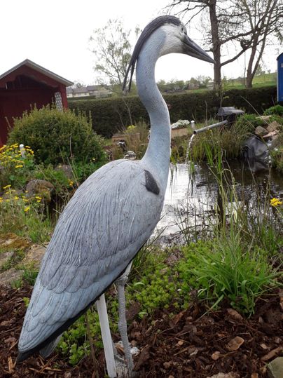A fictional statue of a gray heron stands by a pond. Surrounded by plants and flowers, it gives the garden a natural atmosphere.