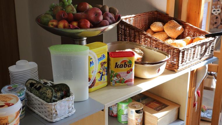 A table with a fruit bowl full of apples and grapes, and a basket of rolls. Next to it is a selection of drinks and food.