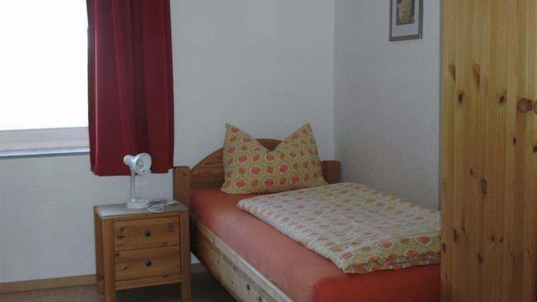 Ein einfaches Schlafzimmer mit einem Einzelbett und bunten Kissen. An der Wand hängt ein Bild und es gibt einen Nachttisch mit einer Lampe.