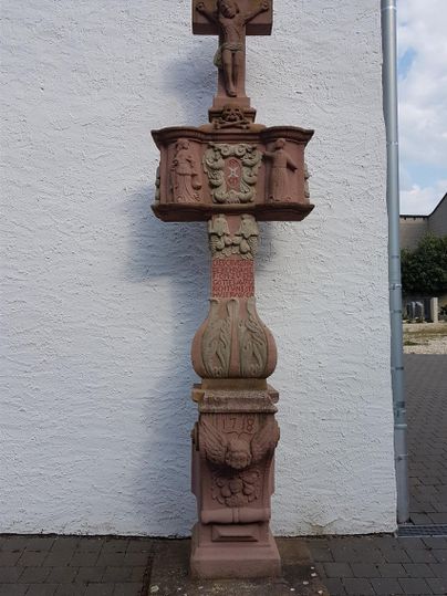 Eine steinerne Statue mit einem Kreuz und Figuren, die auf einem Sockel platziert ist. Der Hintergrund zeigt eine weiße Wand und einen grauen Weg.
