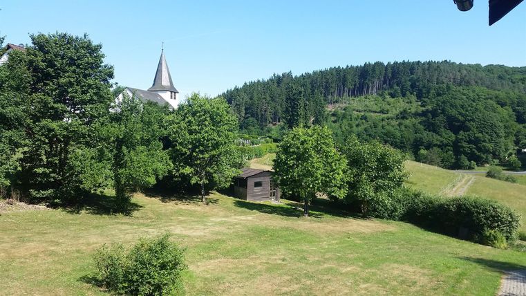 A tranquil landscape with trees and a small cottage. In the background stands a church, and forests surround the scene.