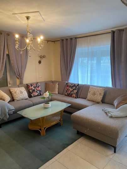 Ein gemütliches Wohnzimmer mit einem großen, grauen Sofa und bunten Kissen. Ein gläserner Couchtisch steht in der Mitte und große Fenster sorgen für viel Licht.