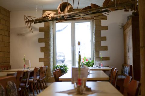 Ein gemütliches Restaurant mit Holztischen und Stühlen. Im Hintergrund sind Pflanzen und ein Fenster mit Blick nach draußen zu sehen.