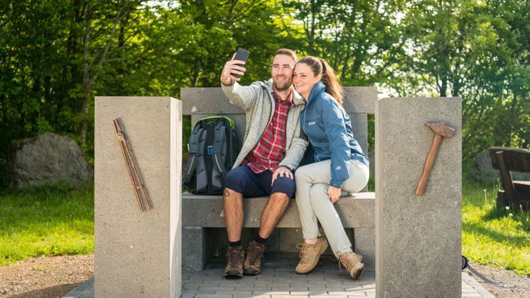 Ein Paar sitzt auf einer steinernen Bank im Freien und macht ein Selfie. Die Bank hat Werkzeuge als Dekoration. Im Hintergrund sind Bäume zu sehen.