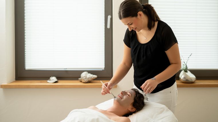 Eine Frau erhält eine Gesichtsbehandlung in einem ruhigen Raum. Die Behandlerin trägt eine schwarze Bluse und appliziert eine Maske auf das Gesicht der Kundin.