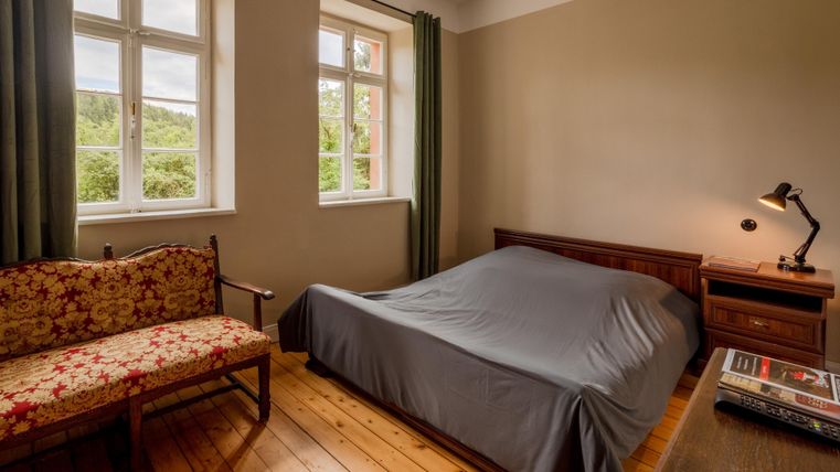 A simple bedroom with a bed and a small table. There are large windows that let in plenty of daylight.