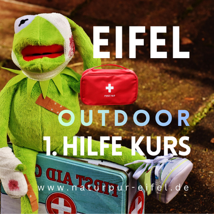 A doll Greenie with a first aid kit sits on a box. The text suggests an outdoor first aid course in the Eifel.