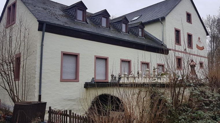 Ein altes, zweigeschossiges Haus mit einem ausgebauten Dachgeschoss. Der Gebäude hat eine helle Fassade und einige Fenster sind mit Jalousien versehen.