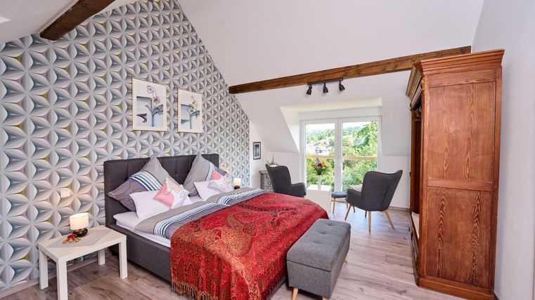 A modern bedroom with a large bed and decorative pillows. The walls are covered with a geometric pattern and there is access to the terrace.