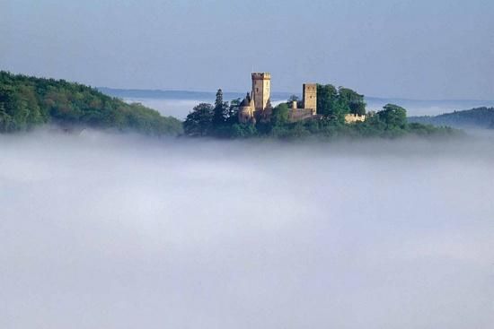 An impressive castle on a hill, surrounded by mist. The sky is clear and the background features gentle hills.