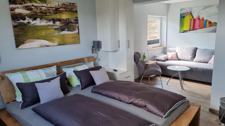 A modern bedroom with two beds and colorful cushions. In the background, there is a living area with a sofa and a table.