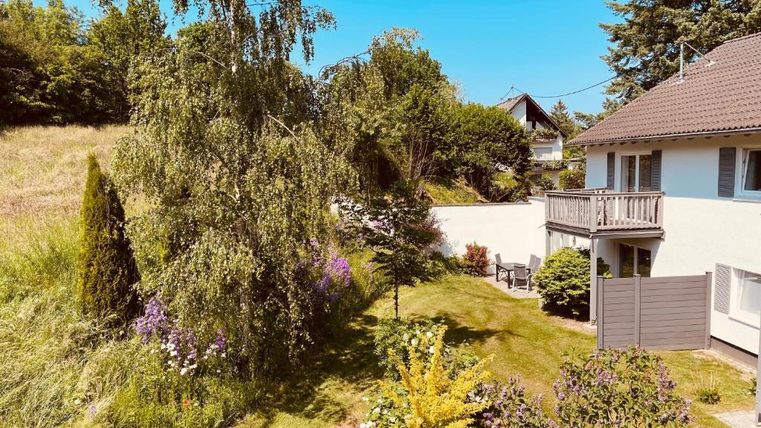 A beautiful garden with different plants and a small pond. In the background, there is a house with a balcony.