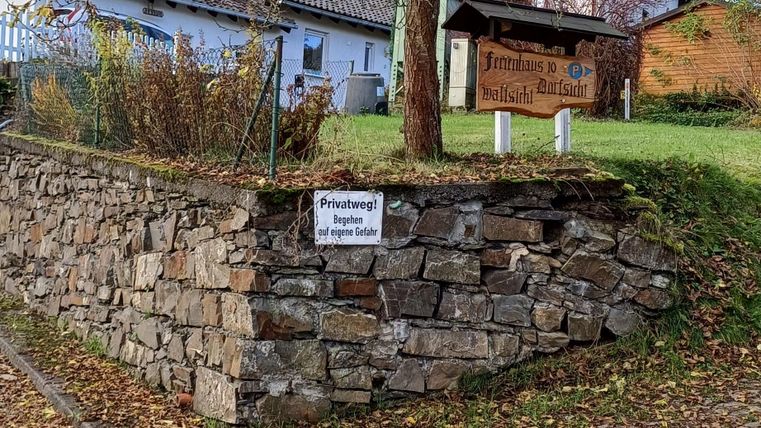 Ein Steinmauer mit einer großen, kahlen Baum und einigen alten Häusern im Hintergrund. Der Boden ist mit Laub und Steinen bedeckt.