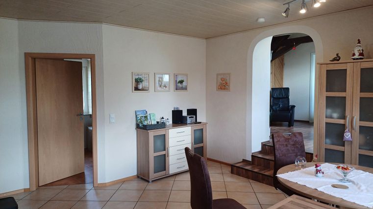A bright and inviting apartment with tiled floors. The room contains furniture, pictures on the wall, and a staircase that leads to another area.