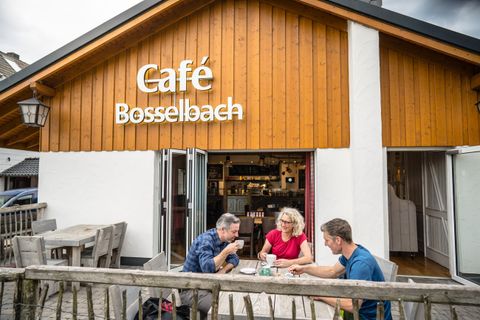 Een gezellig café met de naam "Café Bosselbach". Drie mensen genieten van hun tijd buiten aan een tafel.