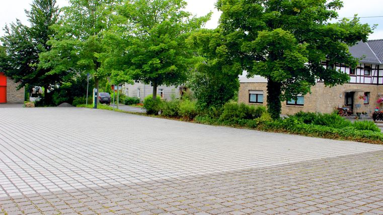 An empty square with a paved floor and some trees. Buildings can be seen in the background.