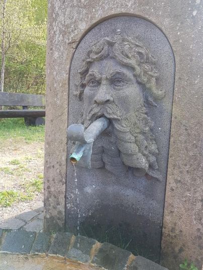 Ein stilisierter Wasserspeier in Form eines alten Mannes mit langem Haar und Bart. Wasser fließt aus seinem Mund in eine kleine Wasserstelle.