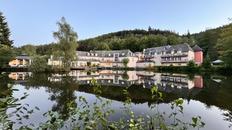 A quiet hotel building is reflected in the clear water of a pond. In the background, green trees and gentle hills are visible.