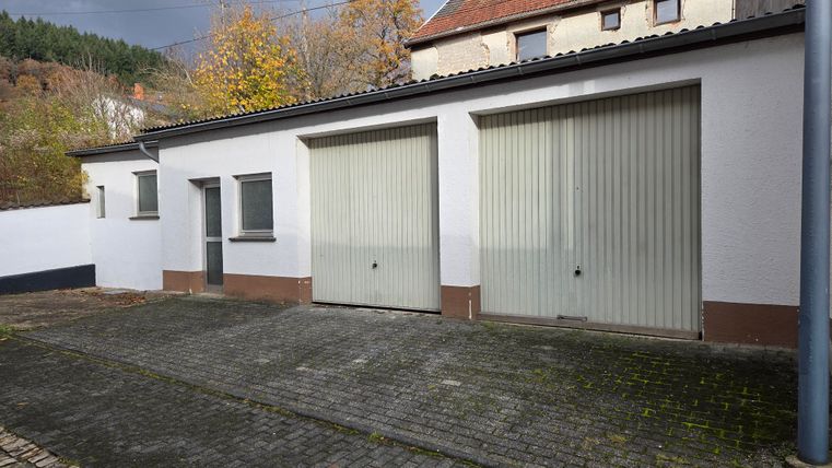 A garage building with two large gray roll-up doors. In the background, trees and another building are visible.