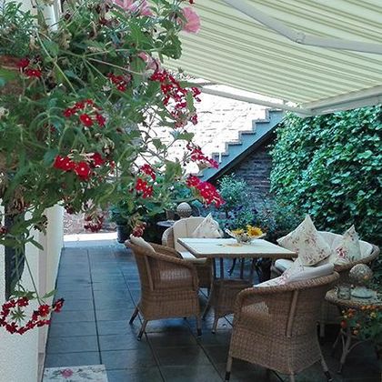 Eine gemütliche Terrasse mit einem Tisch und Stühlen, umgeben von Pflanzen und Blumen. Der Bereich ist gut beschattet und lädt zum Entspannen ein.