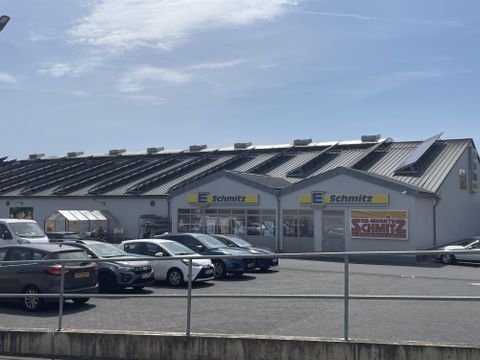 A commercial building named "Schmitz" with a modern roof. In front of the building, several cars are parked on a large parking lot.