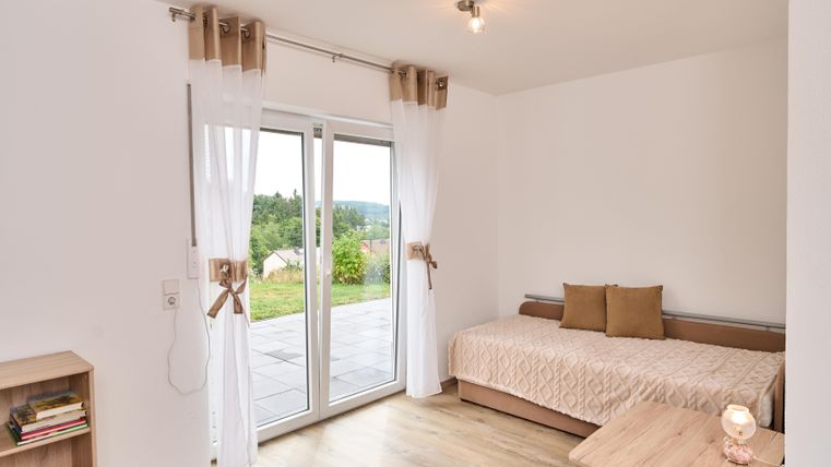 Ein helles Zimmer mit einem Einzelbett und einem kleinen Tisch. Große Fenster bieten einen Blick auf die grüne Landschaft.