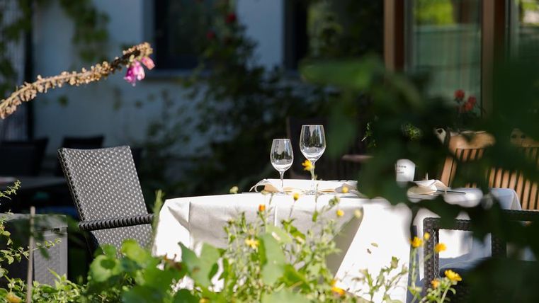 Ein schön gedeckter Tisch im Freien, umgeben von grünen Pflanzen und bunten Blumen. Auf dem Tisch stehen zwei Gläser und einige Geschirrteile.
