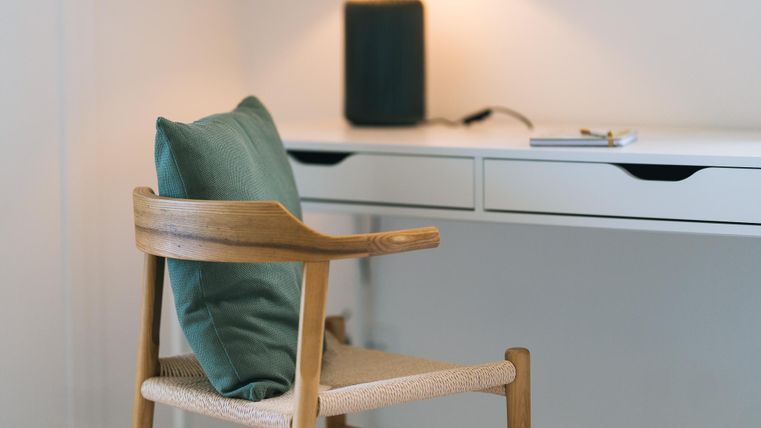 Een moderne bureau met een houten stoel en een kussen. Op de achtergrond zijn een lamp en een luidspreker zichtbaar.