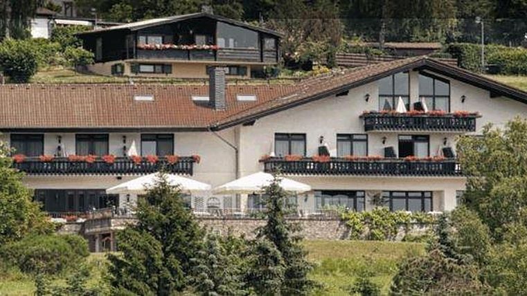 Een charmant hotel met meerdere verdiepingen en balkons, omgeven door groene bomen. Het zonnige buitengedeelte heeft tafels en parasols.