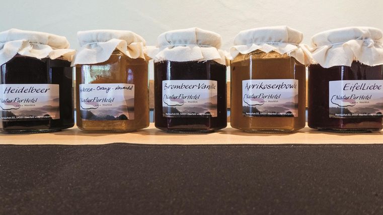 A set of five jars of jam on a table. Each jar has a label with the flavor, including blueberry, strawberry, and several others.