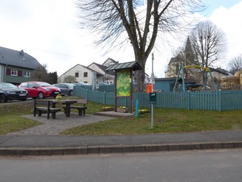Ein kleiner Spielplatz mit einer Schaukel und einem Tischbereich. Im Hintergrund sind ein paar Autos und Häuser sichtbar.
