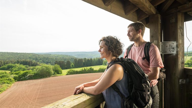 Ein Paar steht auf einer Aussichtsplattform und blickt in die Landschaft. Im Hintergrund sind grüne Wiesen und Wälder zu sehen.