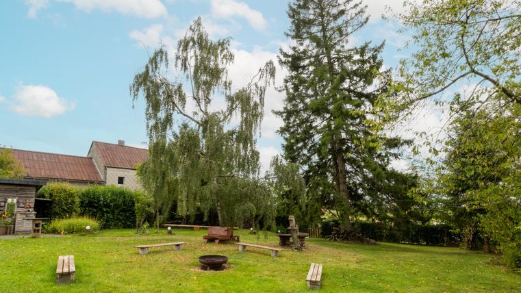 A green meadow with wooden benches and a fire pit in the center. In the background, there are large trees and a house.