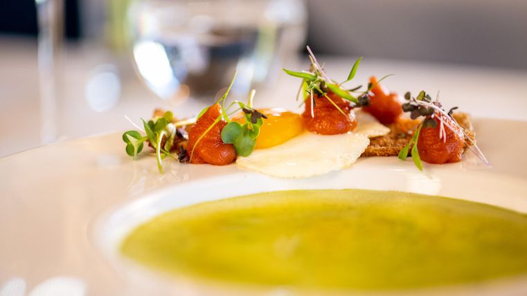 Ein elegant angerichteter Teller mit einer Sauce in Grün und Beilagen aus Ei, Fisch und frischen Kräutern. Im Hintergrund ist ein Glas Wasser sichtbar.
