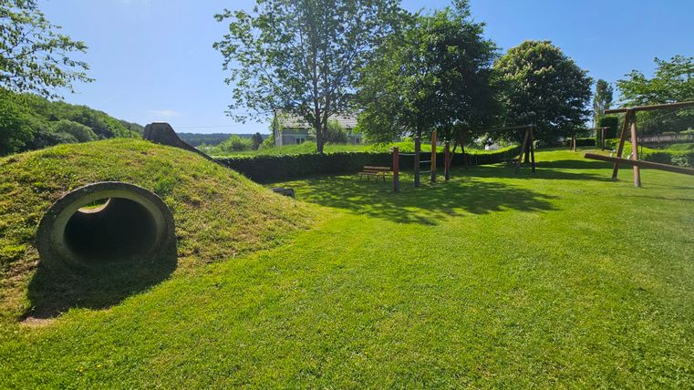 Een grote, groene speelplaats met een heuvelstructuur en een tunnel. Op de achtergrond zijn bomen en speeltoestellen te zien.