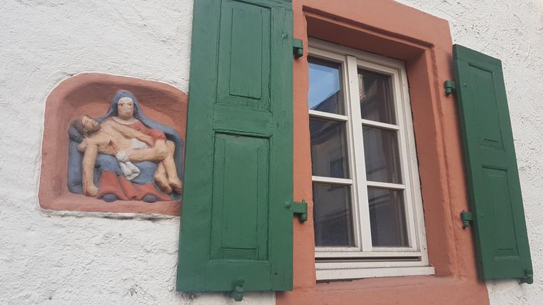 Ein Fenster mit grünen Fensterläden an einer weißen Wand. Daneben ist eine Wandmalerei einer sitzenden Figur zu sehen.