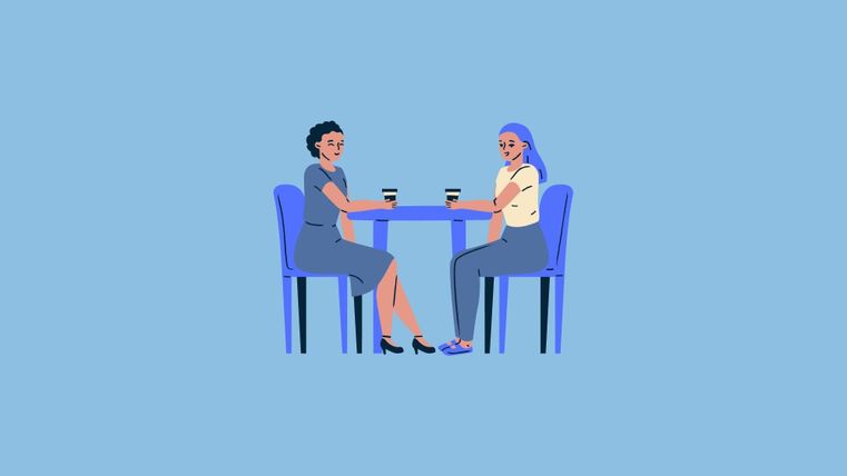 Two women are sitting at a table and having a conversation. Both are holding a cup of coffee in their hands.