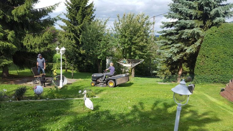 Ein gepflegter Garten mit grünem Gras und hohen Bäumen. Zwei Personen arbeiten mit einem Rasenmäher und einer Schubkarre.
