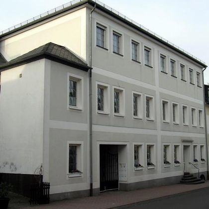 Ein modernes, mehrstöckiges Gebäude mit weißen Wänden und vielen Fenstern. Der Eingang ist klar erkennbar und es gibt einen kleinen Vorgarten.