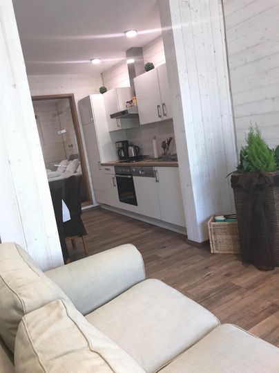 Eine moderne Küchenzeile mit weißen Schränken und Holzfußboden. Im Vordergrund ist ein beiges Sofa sichtbar.