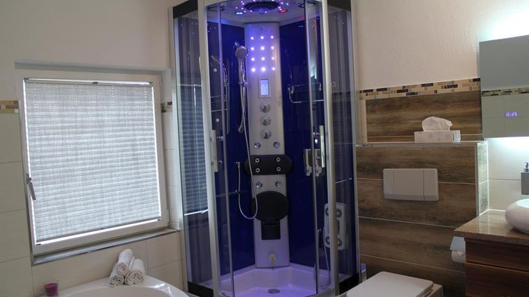 Eine moderne Dusche mit blauem Licht und verschiedenen Funktionen steht in einem stilvollen Badezimmer. Im Hintergrund sind eine Badewanne und ein Waschbecken sichtbar.