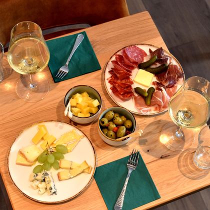 A table with various cheese and cold cuts platters as well as tasty side dishes. There are glasses of white wine to go with it.