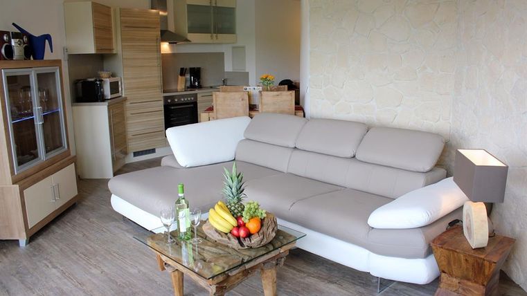 Ein gemütliches Wohnzimmer mit einem modernen, beigen Sofa und einem kleinen Tisch mit frischem Obst. Im Hintergrund ist eine offene Küche mit Holzregalen und modernen Geräten.