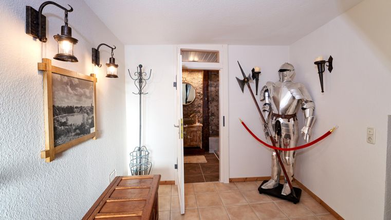 A corridor with a knight in armor and a wooden chest. On the walls, there are wall lights and a painting.
