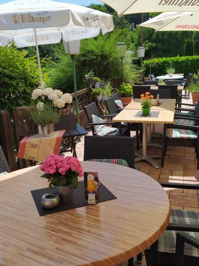 Eine gemütliche Terrasse mit Tischen und Stühlen unter Sonnenschirmen. Blumen dekorieren die Tische und die Umgebung ist grün und einladend.