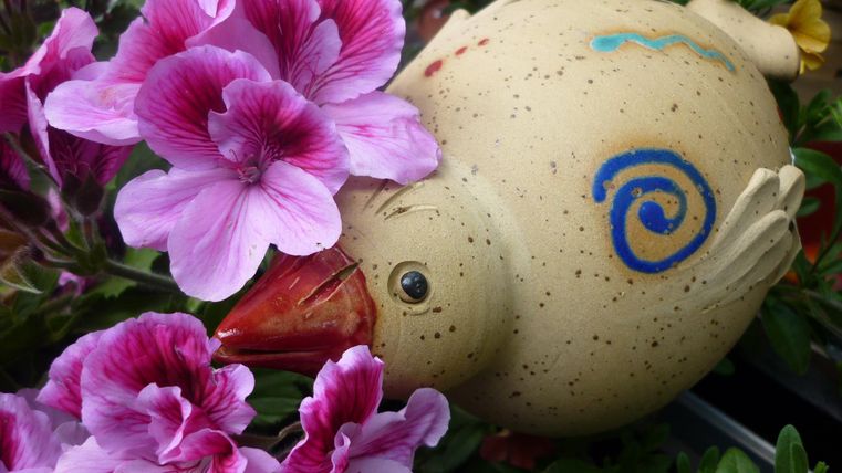 Een kleurrijke, decoratieve kip van keramiek ligt tussen paarse bloemen. De bloemen omringen de kip en geven de scène een vrolijke sfeer.