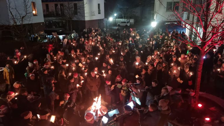 Een grote menigte verzamelt zich op een plein, iedereen met een brandende lantaarn. De sfeer is feestelijk en de omgeving is verlicht door een zwak licht.