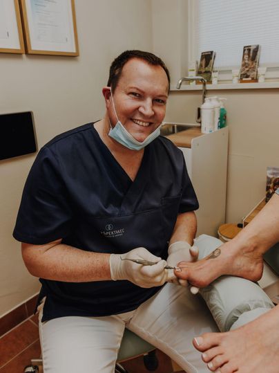 Herr Spengler führt eine Fußbehandlung in seiner Praxis durch. Der Patient wird freundlich betreut, während der Behandler lächelt.