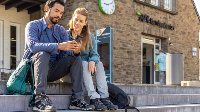 Two people are sitting on the steps in front of a building, looking at a smartphone. In the background, a clock and the logo of a customer service center can be seen.