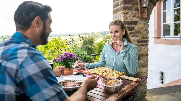 Ein Paar genießt ein gemütliches Essen im Freien. Der Tisch ist mit verschiedenen Gerichten und Blumen dekoriert, mit einer schönen Aussicht im Hintergrund.